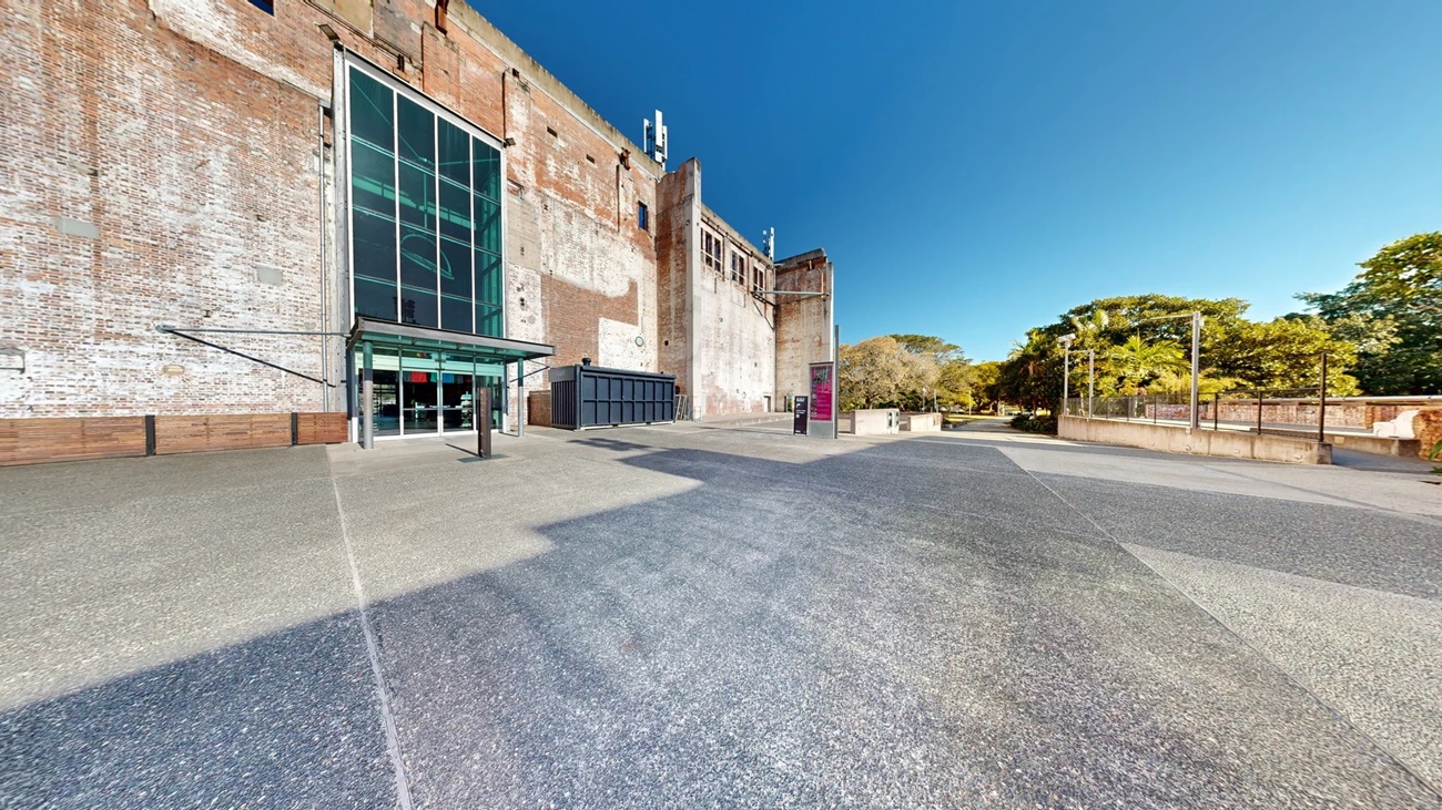 On the left, there is a tall brick building with a large glass entrance and a metal canopy above the doors. In the middle, there is a wide, open concrete area leading to a pink information kiosk. In the background, there are trees and a low stone wall. On the right, there is a continuation of the concrete area with more trees and a railing along the edge. The floor is a mix of smooth and textured concrete.