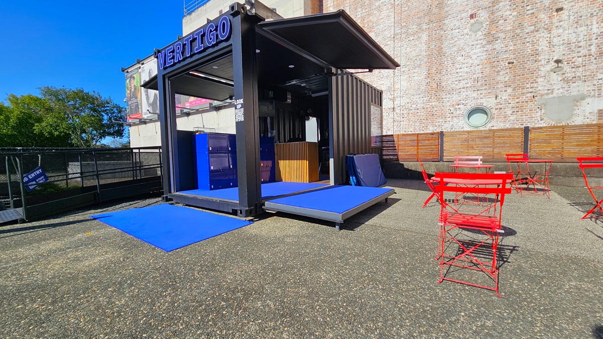 On the left, there is a black metal fence and a tree in the background. In the middle, there is a black structure with a sign reading "VERTIGO" and blue ramps leading up to it. Inside the structure, there is a wooden counter. On the right, there are red metal tables and chairs on a textured concrete floor. The background features a tall brick wall with a circular window and a wooden fence. The floor is textured concrete.