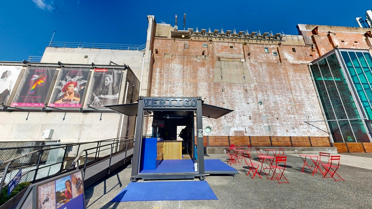 On the left, there is a black metal fence with posters of movies on a white wall. In the middle, there is a black structure with a sign reading "VERTIGO" and blue ramps leading up to it, with a wooden counter inside. In the background, there is a tall, weathered brick wall with various fixtures and a glass section on the right. On the right, there are red metal tables and chairs on a textured concrete floor. The floor is a mix of smooth and textured concrete.
