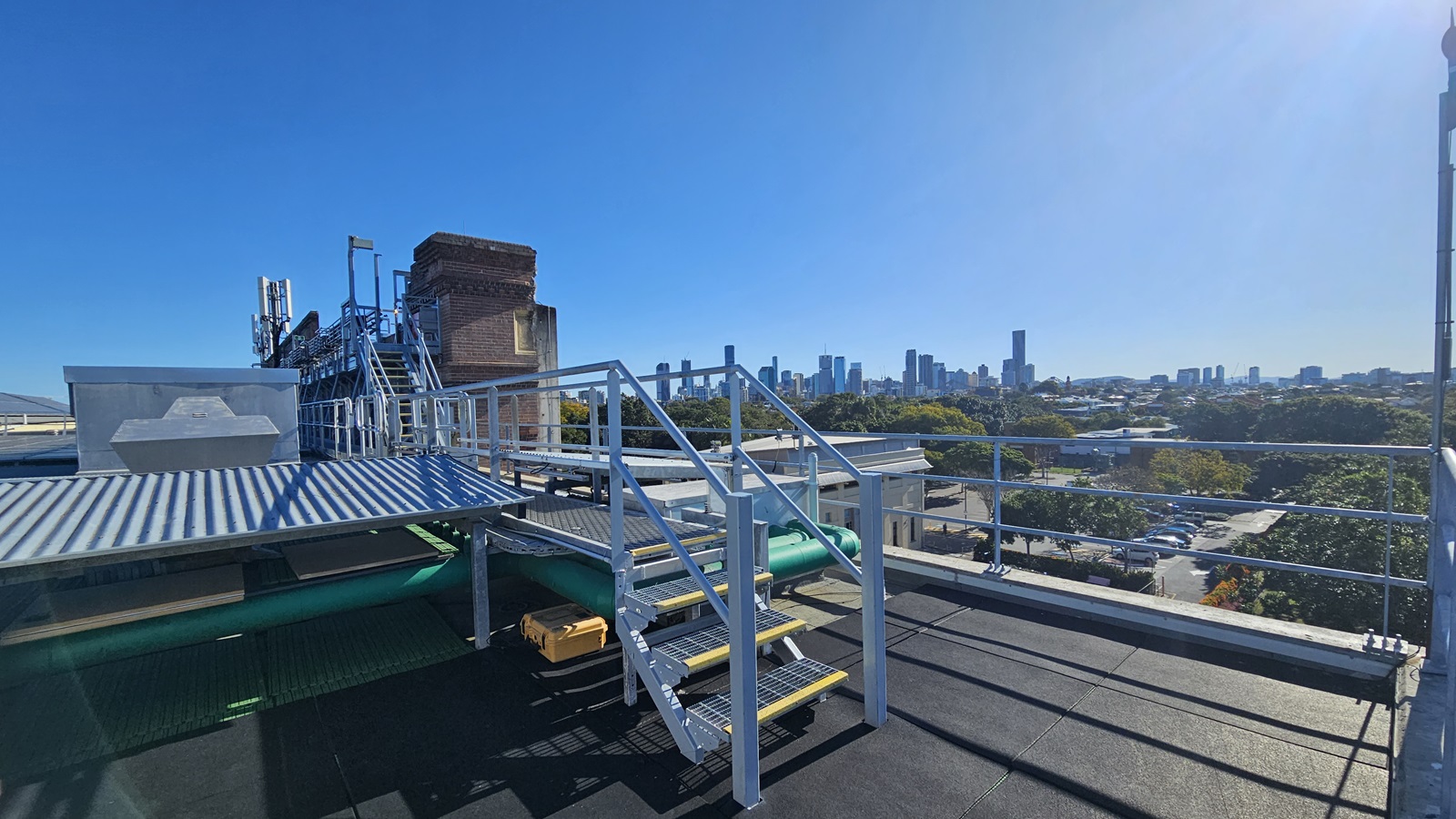 On the left, there is a metal staircase with yellow steps and a handrail, leading up to a platform with a corrugated metal roof. Below the staircase, there are green pipes and a small yellow ladder. In the middle, there is a metal railing and a view of a distant city skyline with tall buildings. On the right, there is a continuation of the metal railing and a view of trees and smaller buildings. The floor is black and appears to be a flat rooftop surface.