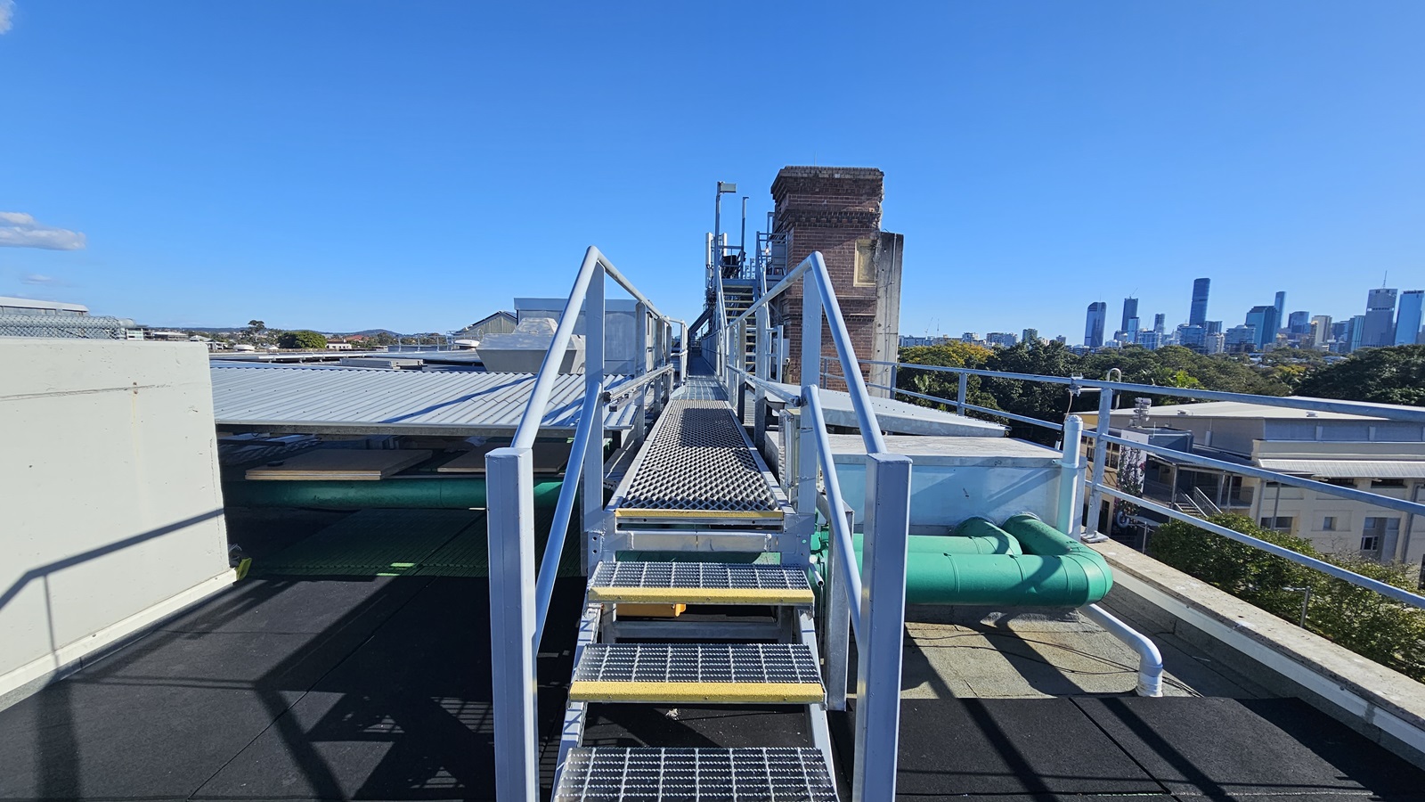 On the left, there is a metal staircase with yellow steps and a handrail, leading up to a platform with a corrugated metal roof. Below the staircase, there are green pipes and a small yellow ladder. In the middle, there is a metal railing and a view of a distant city skyline with tall buildings. On the right, there is a continuation of the metal railing and a view of trees and smaller buildings. The floor is black and appears to be a flat rooftop surface.