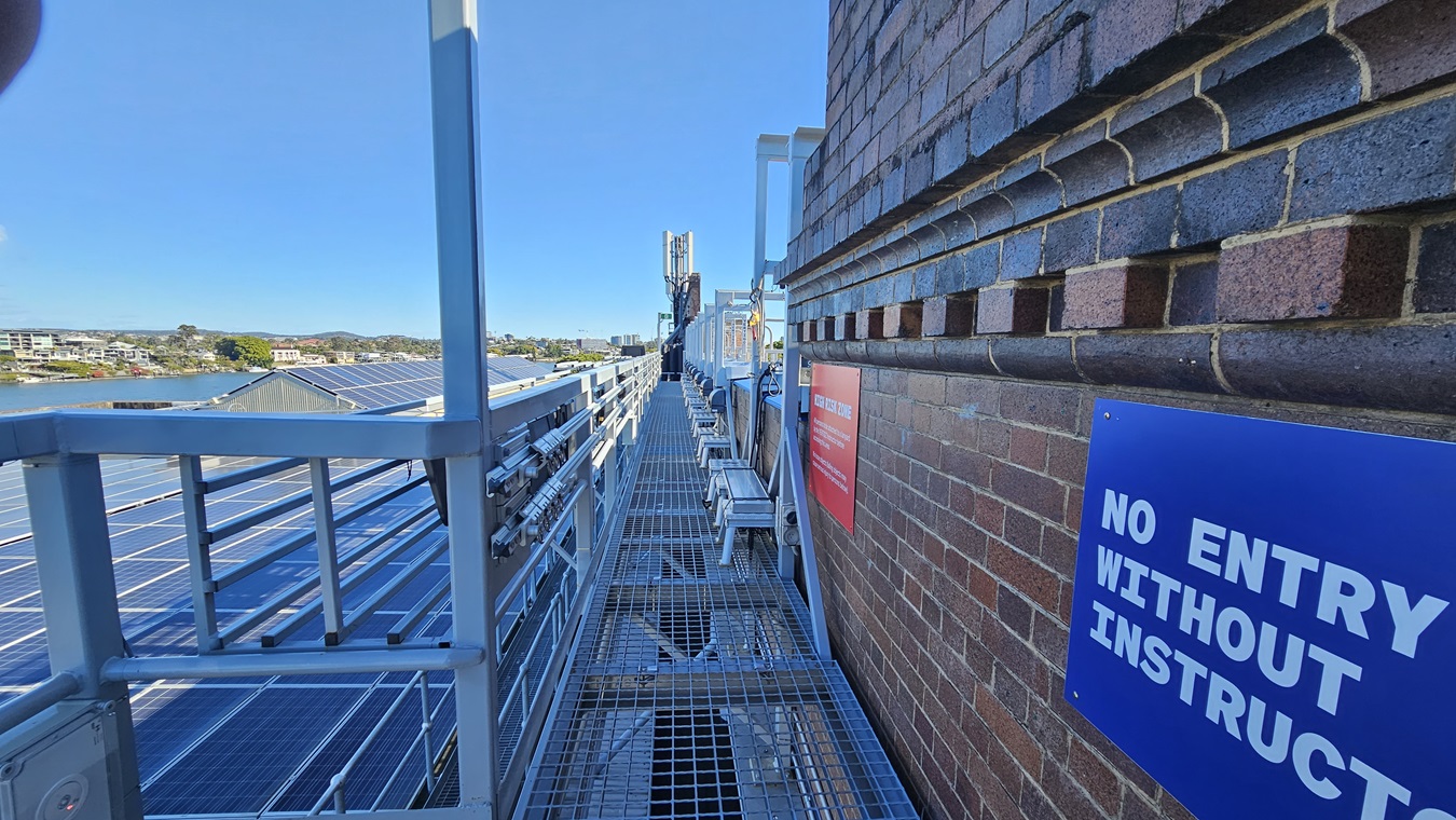 On the left, there is a metal walkway with a railing, running alongside a series of solar panels. In the middle, the walkway continues with metal grating underfoot. In the background, there are distant buildings and a body of water. On the right, there is a brick wall with a blue sign reading "NO ENTRY WITHOUT INSTRUCTOR" and a red sign further along.