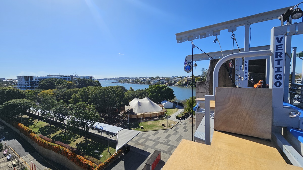 On the left, there is a landscaped area with trees, a pathway, and a low hedge. In the middle, there is a large white tent and a view of a river with buildings on the opposite bank. On the right, there is a wooden platform with metal railings and a vertical sign reading "VERTIGO." The floor is made of wooden planks.