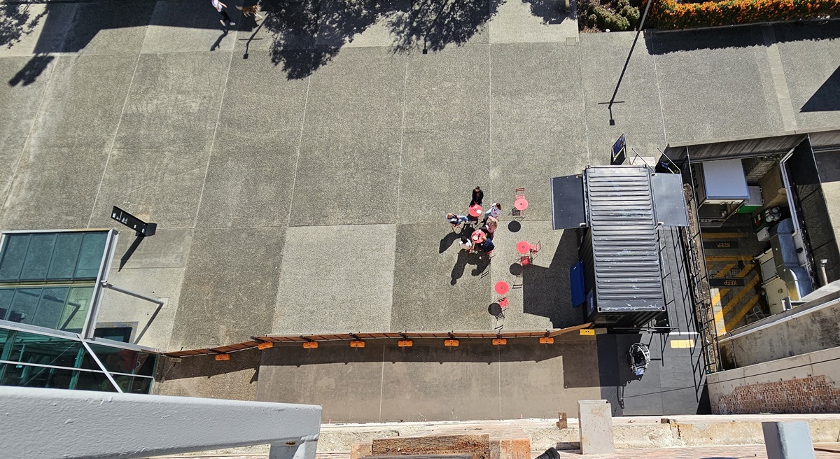 On the left, there is a glass structure with a metal frame and a small signpost. In the middle, there is a group of people standing near red tables with umbrellas, casting shadows on the concrete floor. In the background, there are trees and a pathway. On the right, there is a black shipping container with an open side, revealing equipment inside, and a tall street lamp. The floor is made of large concrete slabs.