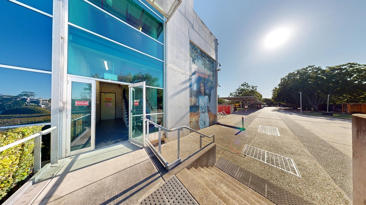 On the left, there is a modern building with large glass windows and an open door with a "No Entry" sign. A metal railing runs along the steps leading up to the entrance. In the middle, there is a wide concrete pathway with metal grates and a painted mural on the building wall. In the background, there are trees and a distant building. On the right, there are more trees and a row of streetlights.