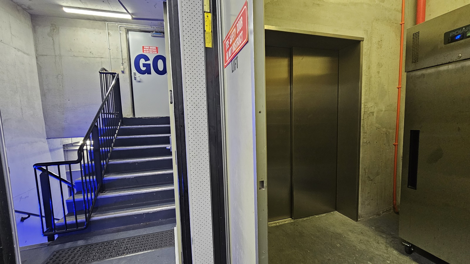 On the left, there is a staircase with black steps and blue lighting underneath, leading up to a white door with a "GO" sign and an emergency exit sign above it. The staircase has black railings and a grey mat at the bottom. In the middle, there is a grey wall. On the right, there is a stainless steel lift with closed doors and a control panel, next to a grey wall with exposed orange piping. The floor is grey concrete, and the ceiling is concrete with fluorescent lighting.