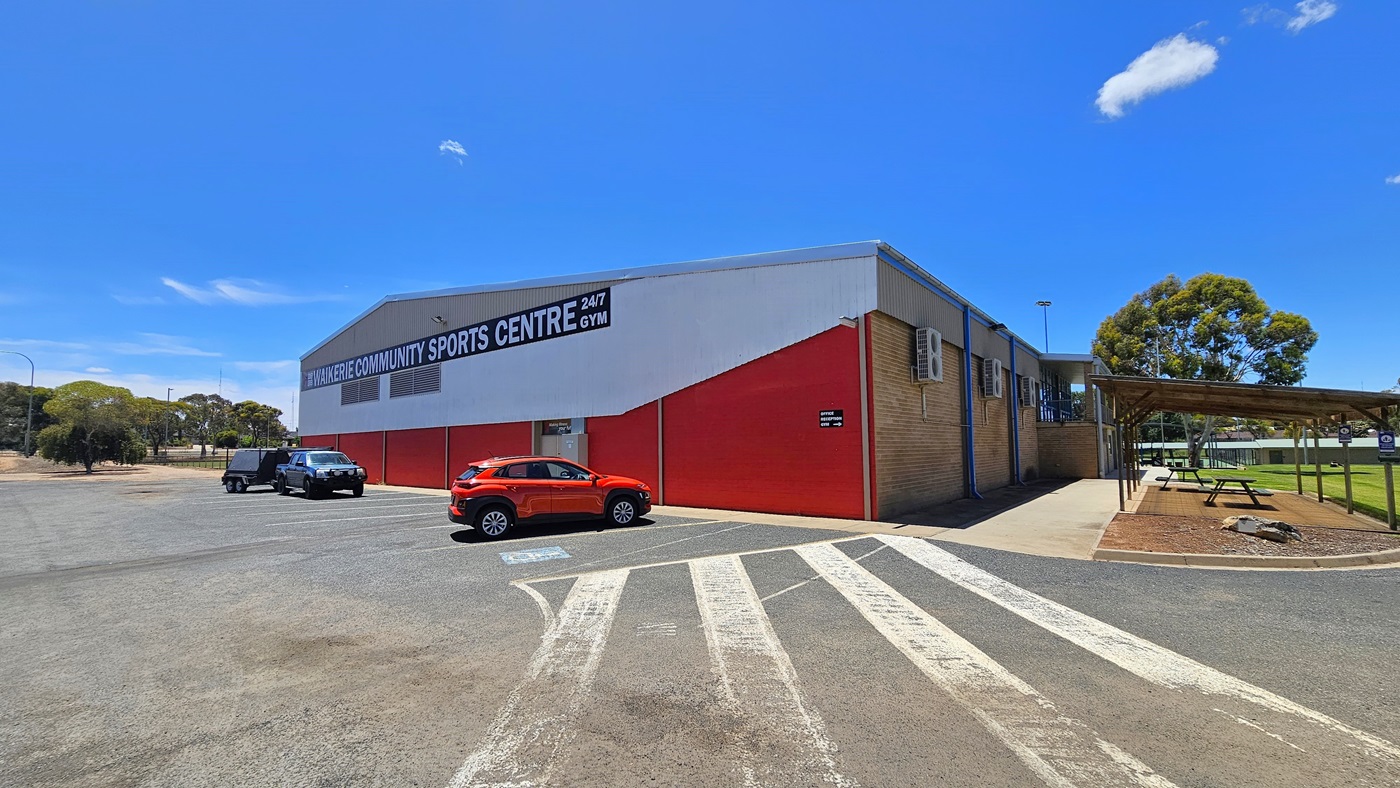 A structure with a red corrugated iron and brown brick exterior stands centrally. Prominent facility signage in bold white letters is displayed. The adjacent concrete car park accommodates three cars of various models and colours, with designated accessible parking on the right, marked by blue wheelchairs and white diagonal lines. To the right of the building is a shaded area with picnic tables, and beyond lies an expanse of green grass. A substantial tree is visible further in.