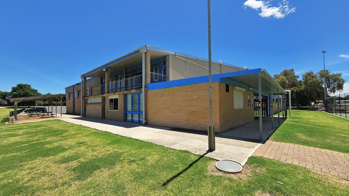 A building with a red corrugated iron and brown brick facade stands centrally. To the left, a shaded area features picnic tables. On the right, a shaded pathway leads to the entrance, and a brick path veers right between the grass. Tall light posts are visible, and a black chain fence surrounds the tennis court.