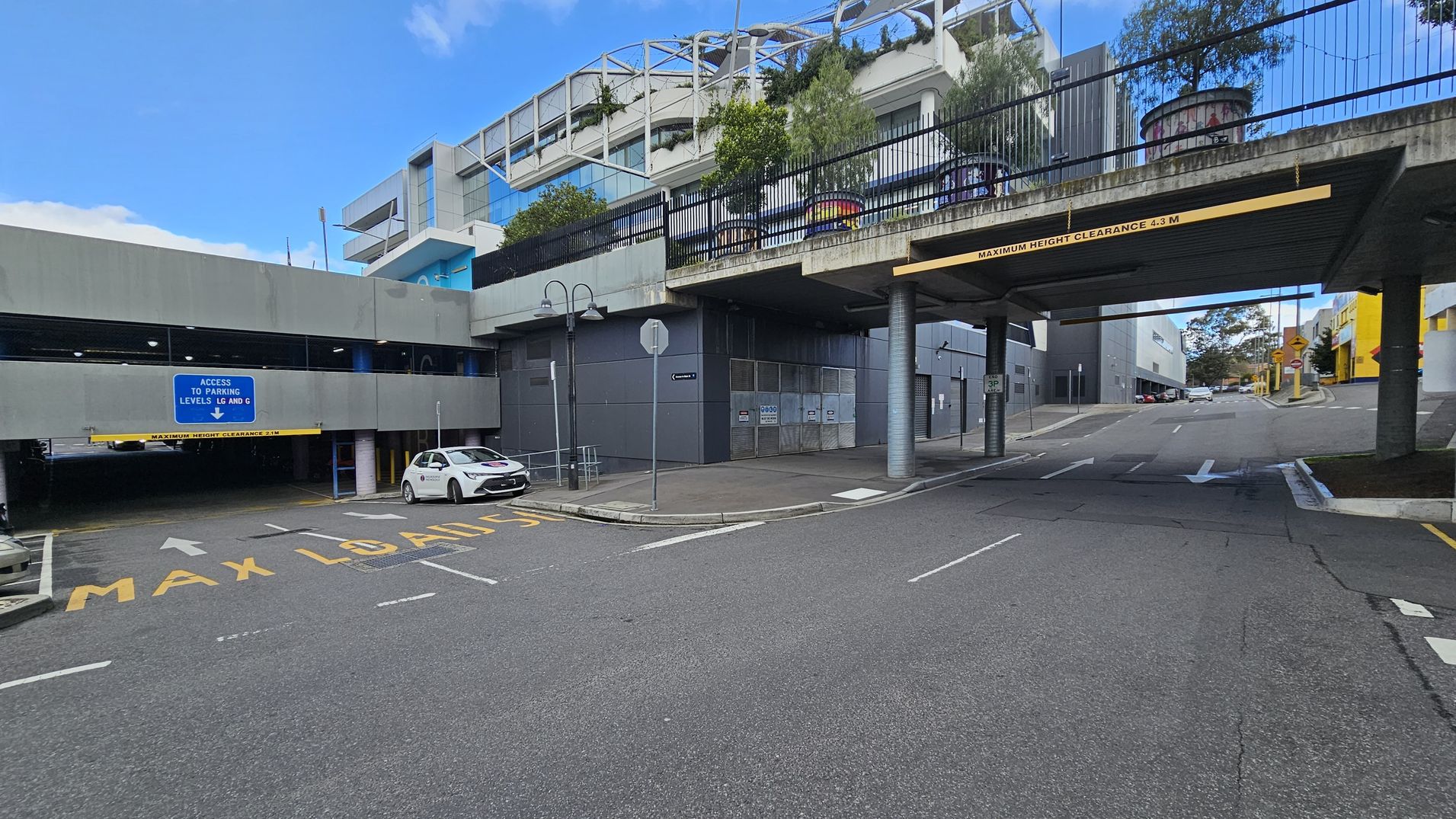 A multi-level car park entrance is on the left, with a blue sign indicating access to parking levels LG and G. A white car is parked nearby. The road has "MAX LOAD 15" painted on it. On the right, a road passes under a bridge with a clearance sign of 4.3 metres. Above the bridge, there are plants and a structure with a metal framework. The background features modern buildings with glass and concrete elements. The road surface is asphalt, and there are directional arrows painted on it.