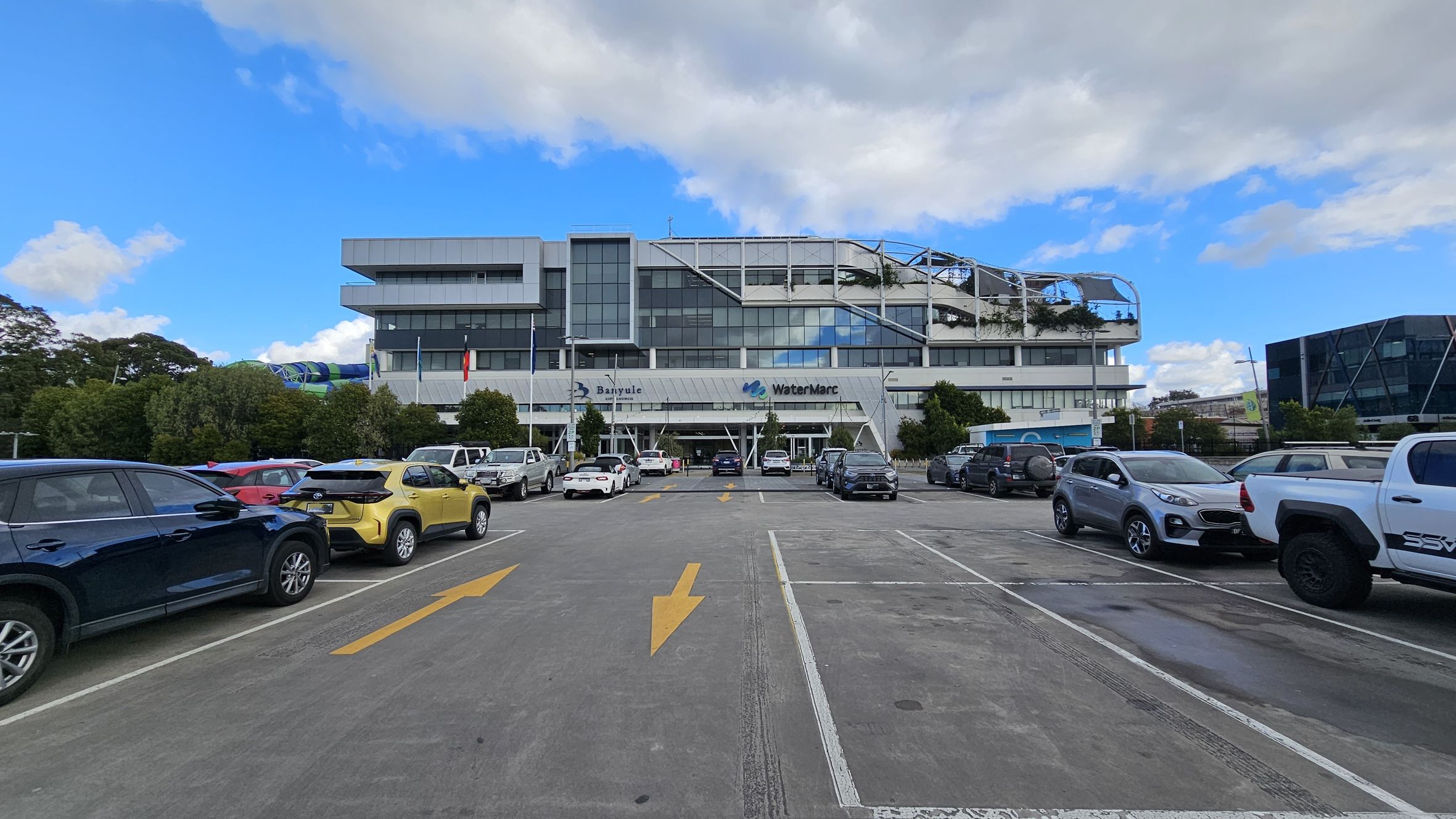 A large, modern building with a glass facade is in the centre, featuring the names "Banyule" and "WaterMarc" on the front. The foreground shows a car park with various vehicles, including a yellow car and a white truck. Yellow directional arrows are painted on the concrete surface. Trees and bushes line the perimeter of the car park, and another building with a geometric design is visible on the right.