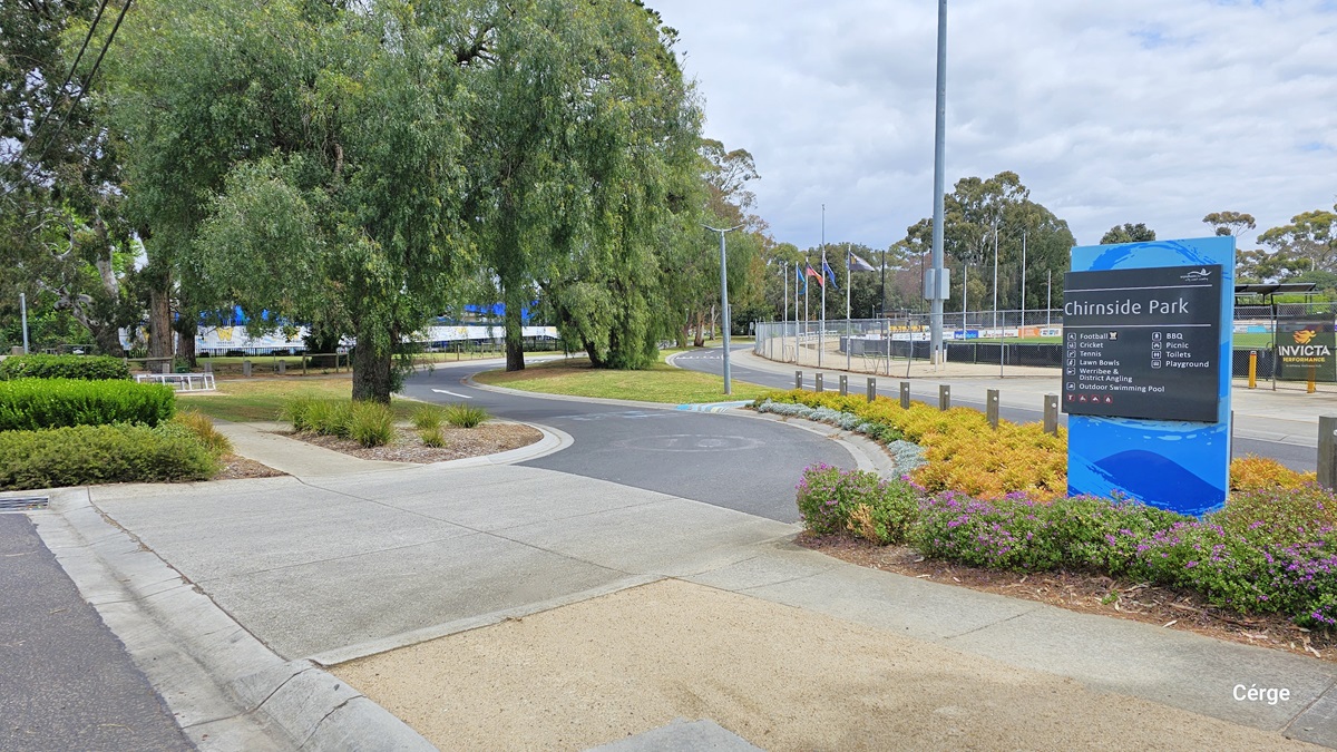 Arrival at Werribee Outdoor Pool via Watton Street, accessible by private vehicles, local buses, and trains. From this entry point, a sidewalk is connected to an asphalt pathway with white directional arrow markings and can be identified by the blue signboard within the garden filled with colorful bushes. The blue signboard shows the list of different amenities at Chirnside Park. The surrounding area has a great landscape filled with tall trees that have wide canopy cover for shade.