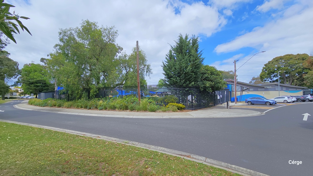 Asphalt pathway with white directional arrow markings leading to the car park at Werribee Outdoor Pool. The facility is enclosed by a tall black iron fence slightly covered by abundant green bushes and different tree types. Tall lamp posts are placed over the parking area where several cars are parked.