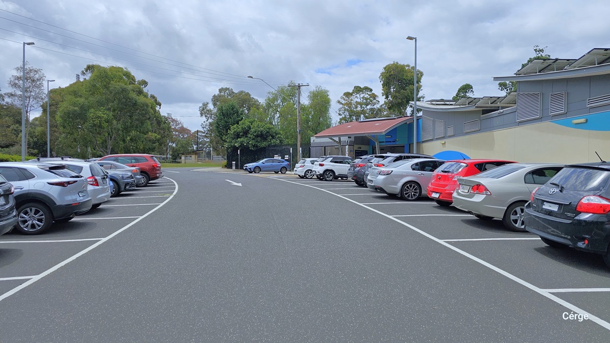 The car parking spaces at Werribee Outdoor Pool are filled with varying types of cars of different colors. The car park is positioned close to the facility and each car parking space has white borderline parking that forms a rectangular pattern. Lamp posts are both present on each side of the pathway. The entrance has a red overhang for shade. The facility is a low-set building with cream, blue, and grey wall accents.
