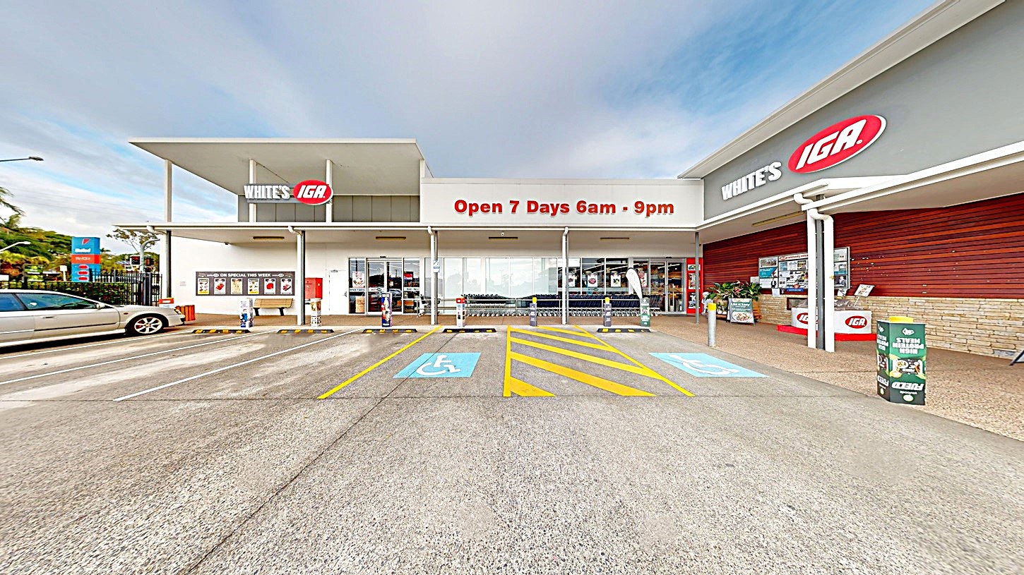 A supermarket entrance with a large sign reading "Open 7 Days 6am - 9pm" in the centre. On the left, there is a row of trolleys and a parked car. On the right, there is a wall with the store's logo and a display of promotional items. The foreground features a concrete floor with marked disabled parking spaces and yellow pedestrian lines. The background includes the store's facade with glass doors and windows.