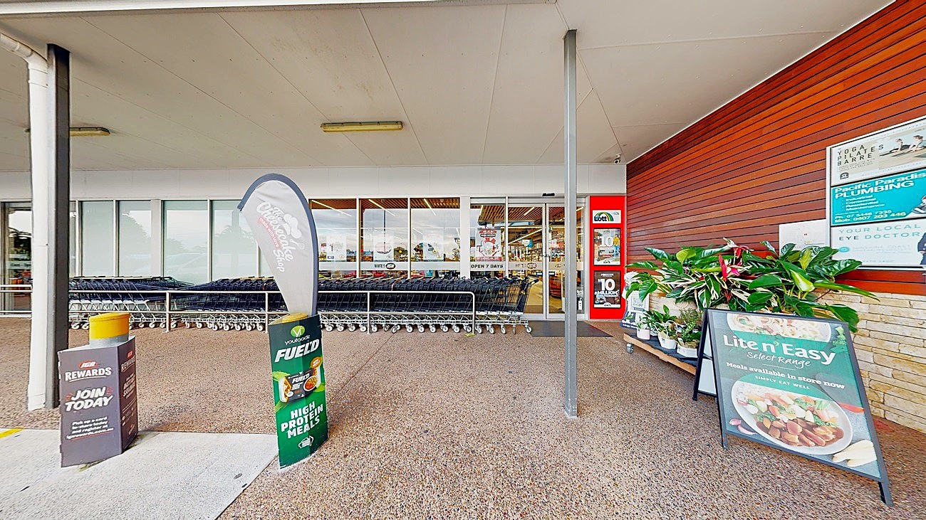 On the left, there is a row of shopping trolleys behind a glass storefront. In the middle, there is a textured concrete floor with a promotional sign for a rewards programme and a flag advertising a product. On the right, there is a wooden wall with posters and a display of potted plants next to a large sign for a meal service. The ceiling is flat and white, extending over the entire area.