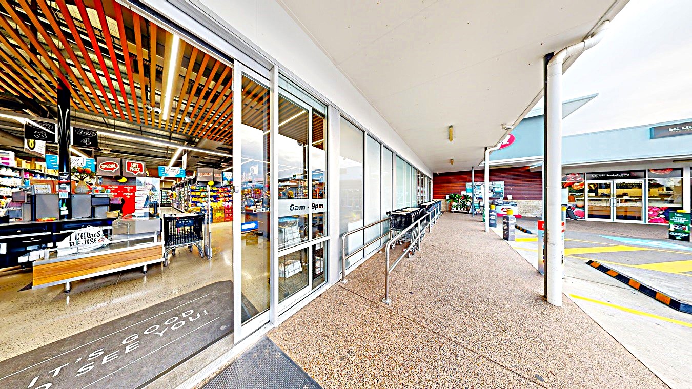 On the left, there is a glass storefront with a sign indicating "6am - 9pm" and a view of the interior with shelves of products. In the middle, there is a walkway with a textured concrete floor and a metal railing. On the right, there is a car park with marked spaces, including yellow pedestrian lines and a few parked cars. The background includes additional store buildings and a covered walkway. The ceiling is flat and white, extending over the walkway area.