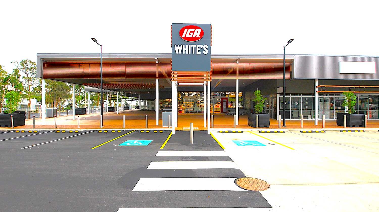 A supermarket entrance with a "WHITE'S IGA" sign in the centre. On the left, there is a covered walkway with wooden slats and a bike rack. On the right, more parking spaces and a glass storefront. In the middle, a pedestrian crossing leads to the entrance. The background features trees and additional buildings. The foreground has an asphalt car park with marked spaces and a puddle.