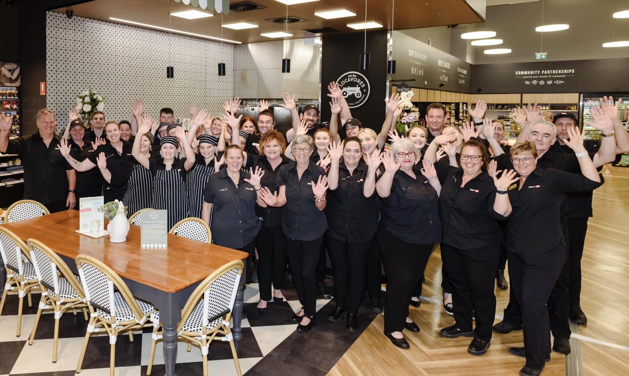 A large group of staff members stands in a café, many raising their hands in a wave. On the left, a man stands casually in a black shirt. The group is predominantly in black attire, with a few wearing striped uniforms. In the foreground, there’s a wooden table with a white vase and menu displayed. The background features a patterned wall, shelves filled with various products, and a menu board. The floor has a black-and-white checkered pattern, while the ceiling has modern light fixtures.