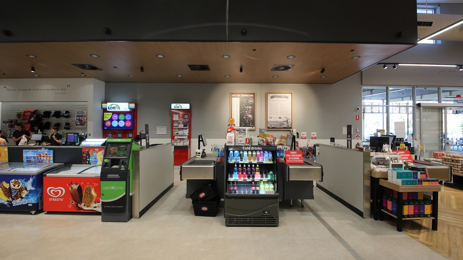 A direct view of three cash register aisles, with two ice cream freezers situated to the left next to an ATM and marketing materials for ‘Lotto’ can be seen towards the back. To the left three people can be seen chatting near the express check out area. To the right is a small display featuring boxes in a range of colours including green, yellow and orange. Directly ahead is a small drinks fridge and on the far back wall are two large prints with brown frames.