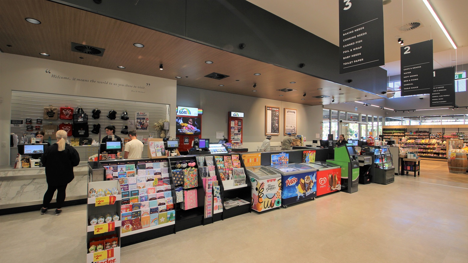 Directly ahead to the left is a lady dressed in black being served by another lady at the express check out area. On the back wall is a range of bags and other merchandise as well as marketing materials for ‘Lotto’ on display to the right. In front of this express checkout area is a range of chocolates, a display of gift cards, wrapping paper and ribbons next to the newspaper and magazine stand. Next to three ice cream freezers and an ATM leading up to the three cash register aisles.