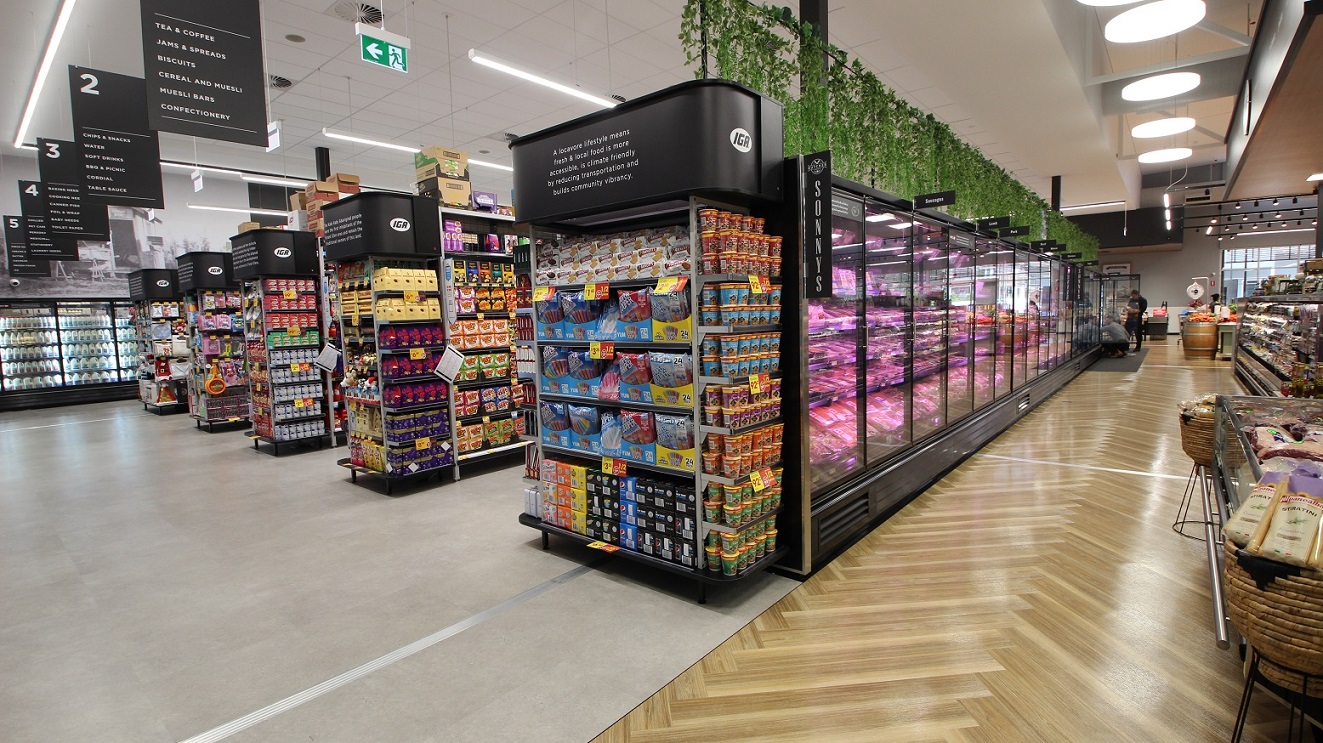 A row of five grocery aisles are to the right featuring a fridge full of dairy products at the far end. Each aisle has a sign at the end listing a number of the products available in that aise. The aisle closest to the front features a range of meat products and there is green, plastic foliage hanging from black wire above the meat fridge. Across from the meat fridge is a range of condiments and other specialty items on display.