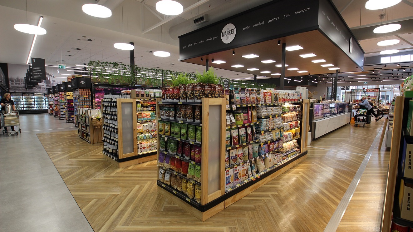 To the far end is the dairy fridge where a worker can be seen with stock, situated across from the five grocery aisles. In the centre of this image is the ‘Market Deli’ which is a square shaped area with large black signage above it. Adjacent to this are two sections of wooden shelving that features items such as cake mixes, mueslis and other dry goods. Green plastic foliage is used throughout. Further to the right is a customer with a shopping trolley.
