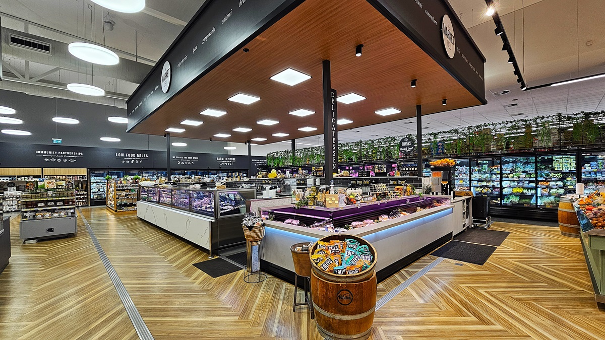 A supermarket interior with a wooden chevron-patterned floor. On the left, there are shelves with various packaged goods. In the middle, there is a large island with refrigerated display cases containing meats and cheeses. The ceiling above the island is wooden with square lights. On the right, there are refrigerated sections with glass doors displaying dairy products and vegetables. The background features more shelves and a section with potted plants on top.