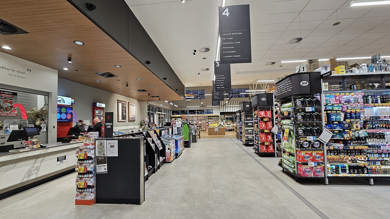 A supermarket aisle with a light grey tiled floor. On the left, there is a customer service counter with staff and a red vending machine. In the middle, there are self-checkout stations. The ceiling above is a mix of wooden panels and white tiles with recessed lighting. On the right, there are shelves stocked with various beverages and snacks. The background features more aisles and a large sign indicating different sections of the store.