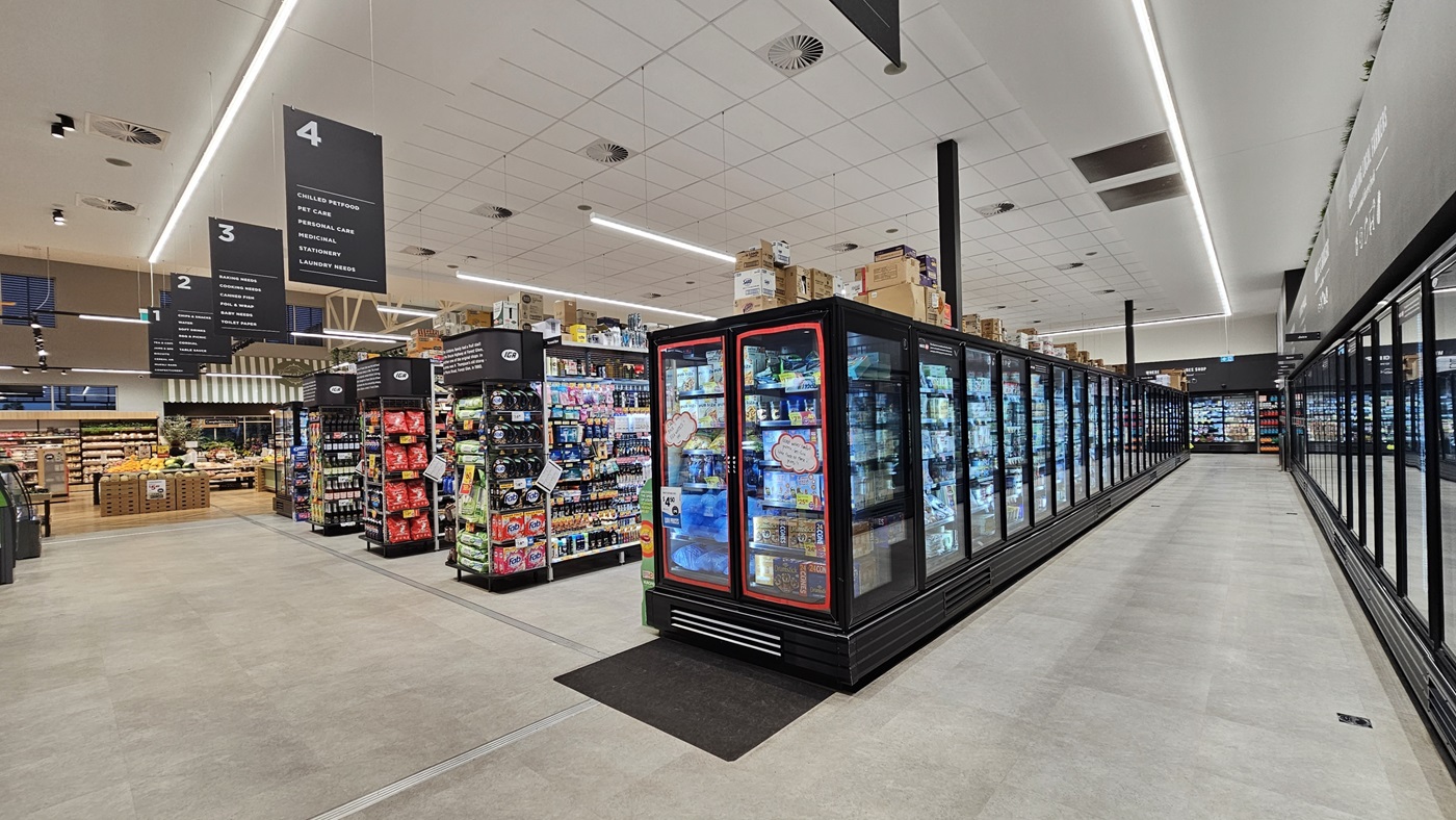A supermarket interior with a light grey tiled floor. On the left, there are shelves stocked with snacks, drinks, and household items. In the middle, there is a long row of refrigerated display cases filled with frozen foods. The ceiling is white with recessed lighting and ventilation ducts. On the right, there are more refrigerated sections with glass doors containing dairy products and beverages. The background features additional aisles and a section with fresh produce.