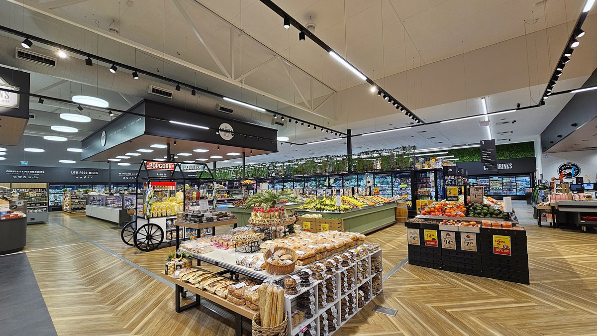 A supermarket with a wooden chevron-patterned floor. On the left, there are shelves with baked goods and a display of fresh produce. In the middle, there is a large island with various fruits and vegetables. The ceiling is high with white panels and black track lighting. On the right, there are refrigerated sections with glass doors containing dairy products and meats. The background features more produce displays and a section with potted plants on top of shelves.