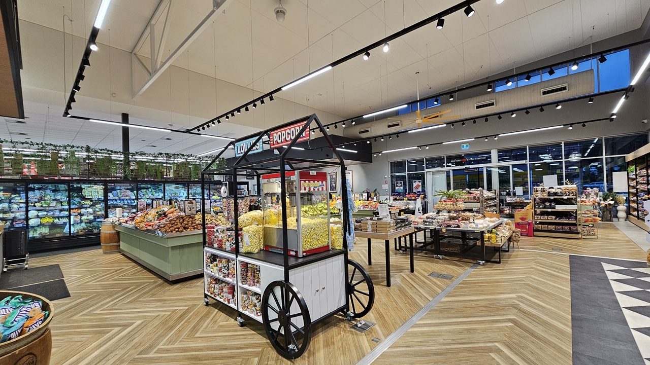 A supermarket with a wooden chevron-patterned floor. On the left, there are refrigerated sections with glass doors displaying fresh produce and a green display table with fruits and vegetables. In the middle, there is a black popcorn cart with a red sign and a variety of snacks. The ceiling is high with white panels and black track lighting. On the right, there are more display tables with baked goods and packaged items, and a section with potted plants on top of shelves.