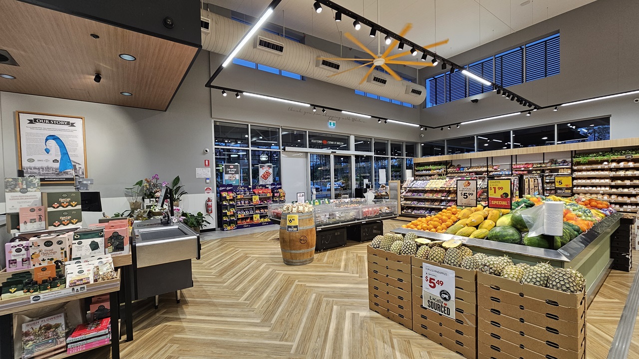 A supermarket with a wooden chevron-patterned floor. On the left, there is a display of chocolates and a small sink area. In the middle, there is a wooden barrel and a refrigerated section with packaged meats. The ceiling is high with white panels, black track lighting, and a large yellow fan. On the right, there are wooden display tables with various fruits and vegetables, including watermelons and pineapples. The background features large windows and more shelves with produce.