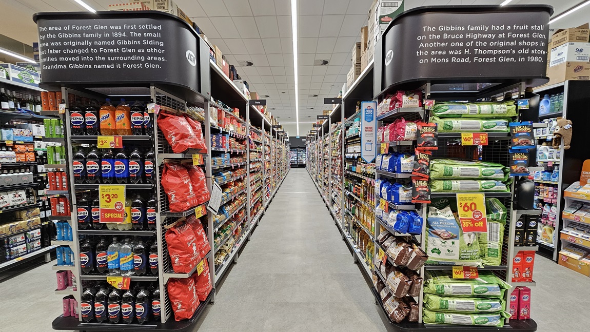 A supermarket aisle with a grey tiled floor. On the left, there are shelves stocked with various snacks, including crisps and soft drinks. On the right, there are shelves filled with pet food and cleaning supplies. In the middle, the aisle extends towards the back of the store, with overhead signs providing information about the history of the area. The ceiling is white with rectangular fluorescent lights. The background features more aisles and a checkout area.