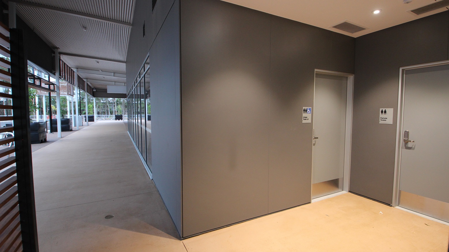 To the left of the picture is a view of the car park featuring the pot plants. To the right are grey walls and a beige floor leading into the female toilets and the Unisex accessible toilet. Both of these doors are light grey in colour and feature silver door handles with a pass code entry.