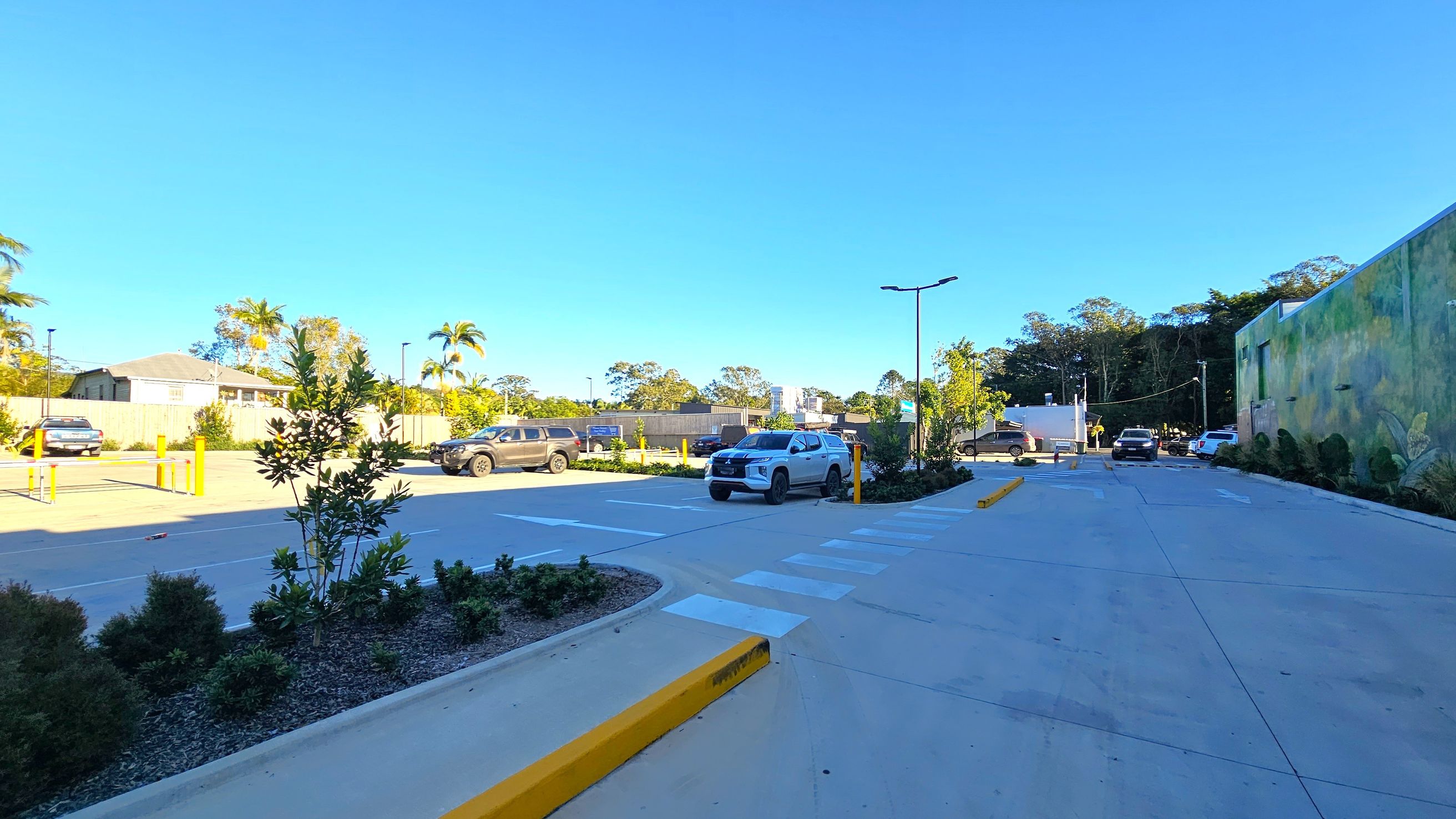 A car park with several vehicles is visible. On the left, there are parked cars and a small garden area with shrubs and a tree. On the right, a large wall with a mural and more parked cars. The middle features a pedestrian crossing and a clear road leading further into the parking area. The background includes trees and a few buildings. The ground is paved, and there are streetlights along the road.