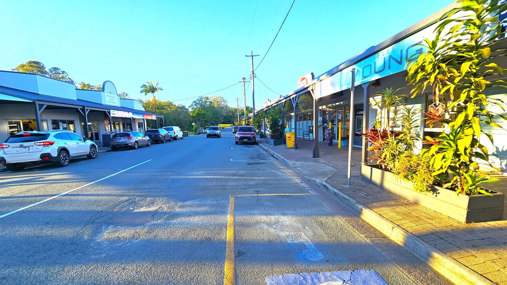 A street scene with shops on both sides. On the left, several cars are parked in front of a row of shops with a covered walkway. On the right, more shops with a similar covered walkway, featuring large potted plants. The road is lined with parked cars and a few utility poles with overhead wires. The background shows trees and greenery. The pavement is a mix of asphalt and brick, and the lighting suggests late afternoon or early morning.