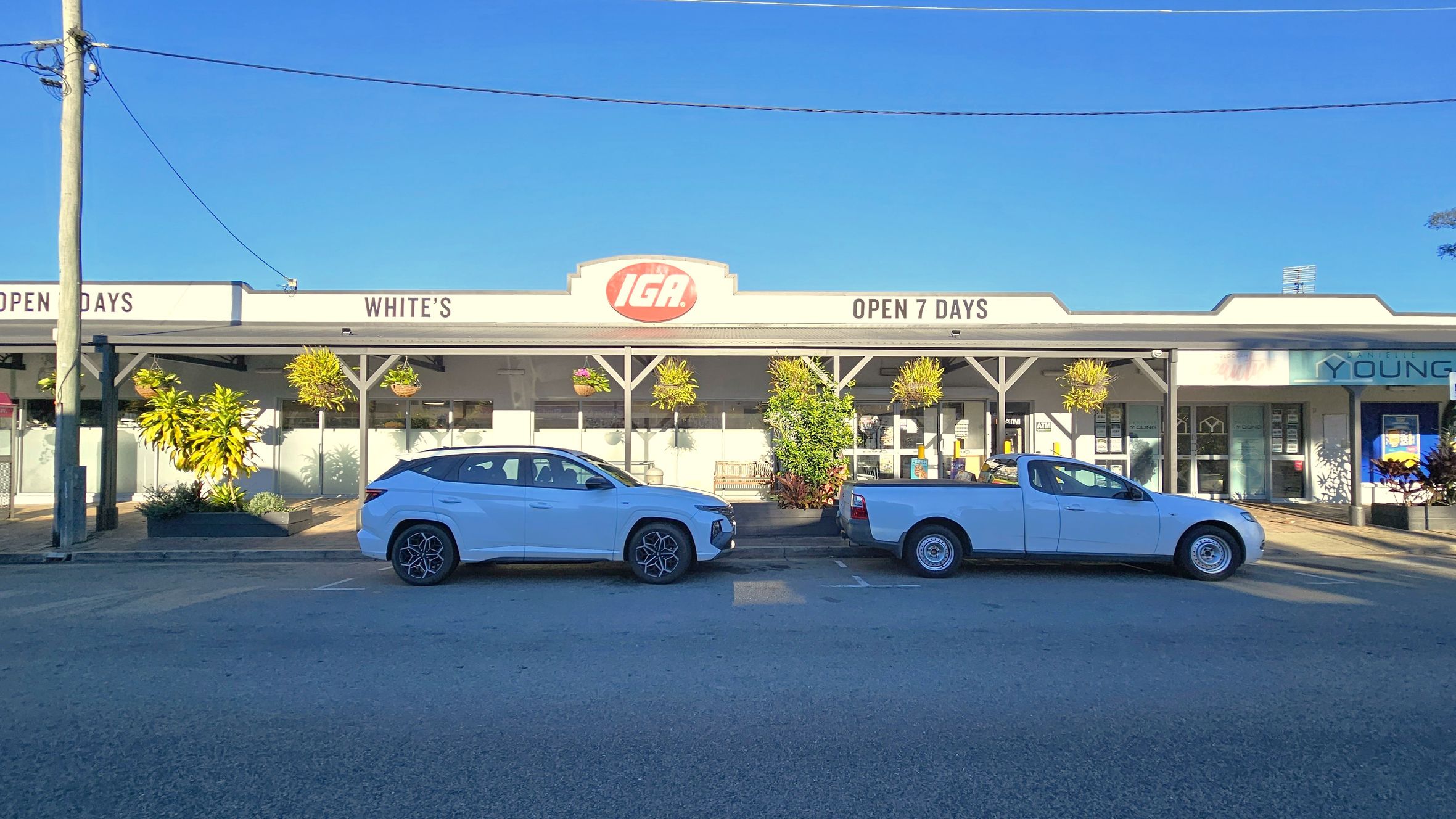 A storefront with a sign reading "IGA" and "Open 7 Days" is in the centre. The building has a grey roof and several hanging plants. In front, a white SUV and a white pickup truck are parked on the street. To the right, there's a sign for "Young" on a neighbouring shop. A power pole with wires is on the left. The pavement is visible in the foreground.