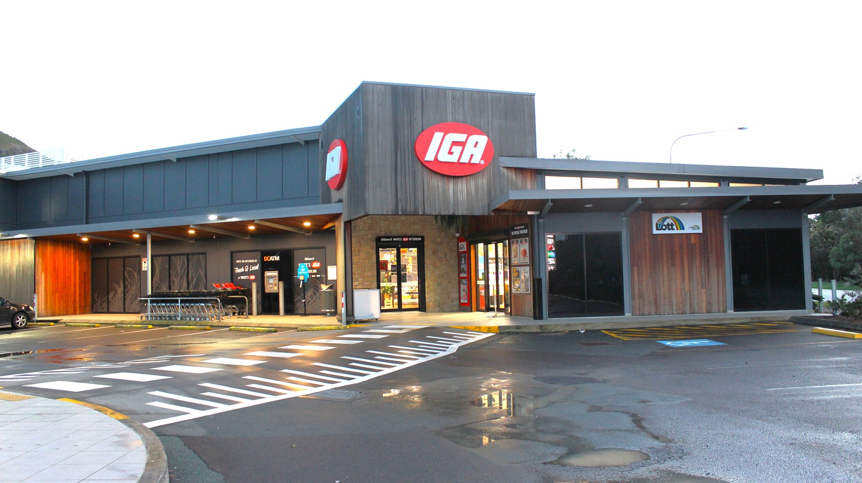White's IGA Mt Coolum has a red oval-shaped sign with white IGA letters located on the façade. The building design is modern and is painted with grey and brown. There are shopping trolleys available outside the store and an ATM. There are also several parking spaces in front of the building indicated by painted white lines that served as space partitions for each car. There is an accessible car parking space available directly outside the store.