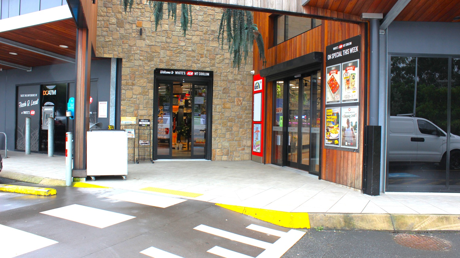 To the left, there are grey poles and a cash machine marked as "DCATM" next to a small shopping cart corral. The path leads to a slightly elevated concrete area where a white storage unit is positioned. On the right side, two large glass doors open into a store labelled "WHITES MT COOLOM", with promotional signs displayed next to them. The floor is a mixture of concrete and tiles with yellow and white lines marking pedestrian crossings.