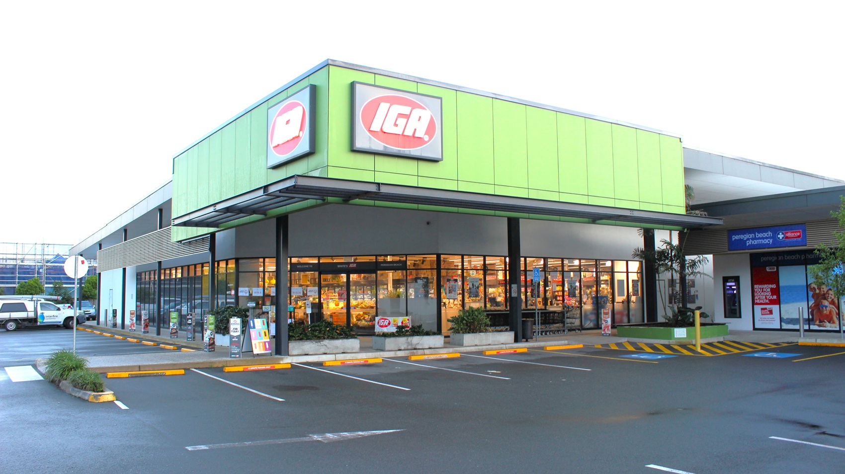 An outside look at the sophisticated IGA shopping building. The top of the building has green walls with the IGA brand name, and half of the wall of the shopping building is made of glass. Two accessible car spaces are positioned toward the right of the store entrance.