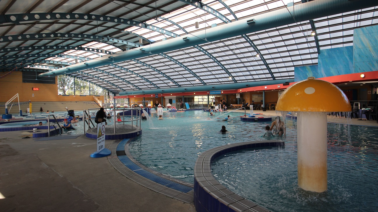 Adults and children are enjoying their bonding time at the children’s pool. Aside from the mushroom fountain, there are metal tubes spurting a standard flow of water on the edges of the pool. A no-diving sign is also placed to caution all the guests. Visible further to the back of the image is a digital clock and a white hoist sling for lifting persons with mobility disabilities.