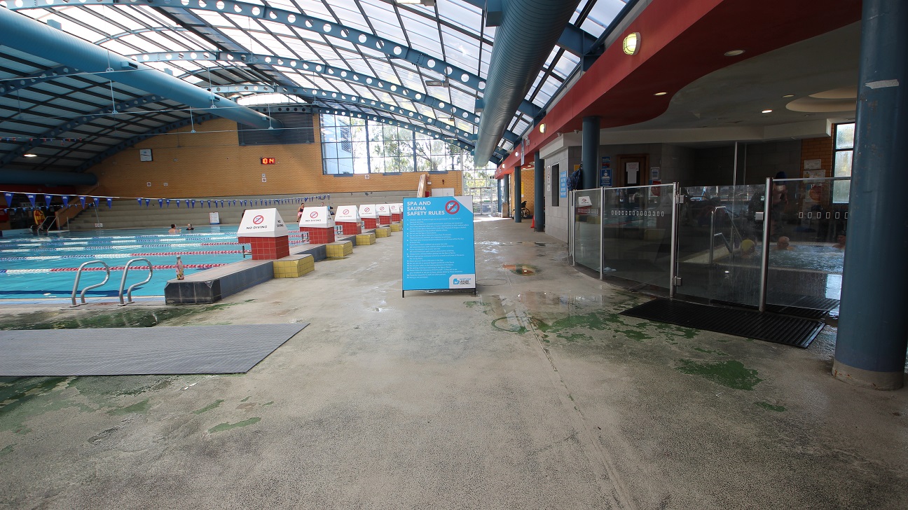 The image shows the 8-lane 25m lap pool on the left side while the jacuzzi can be found on the right side, separated by the hip-length glass walls. The lanes are separated by white, red, and blue floating lane ropes. Small triangular flags placed horizontally in the pool area can be seen hanging 5 meters from the water's surface. On the red elevated section, the right side of the pool, are triangular white signboards cautioning swimmers that no diving is allowed.