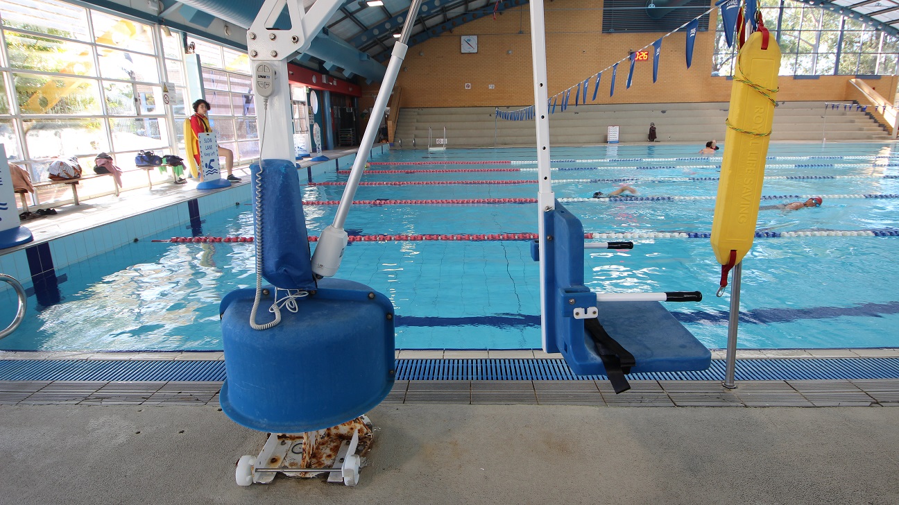 White pool hoist with blue seating chair at the 8-lane 25m lap pool. Near it is a yellow oblong floating platform used by lifeguards when saving someone. Long benches are placed against the glass walls for bags and other personal belongings.