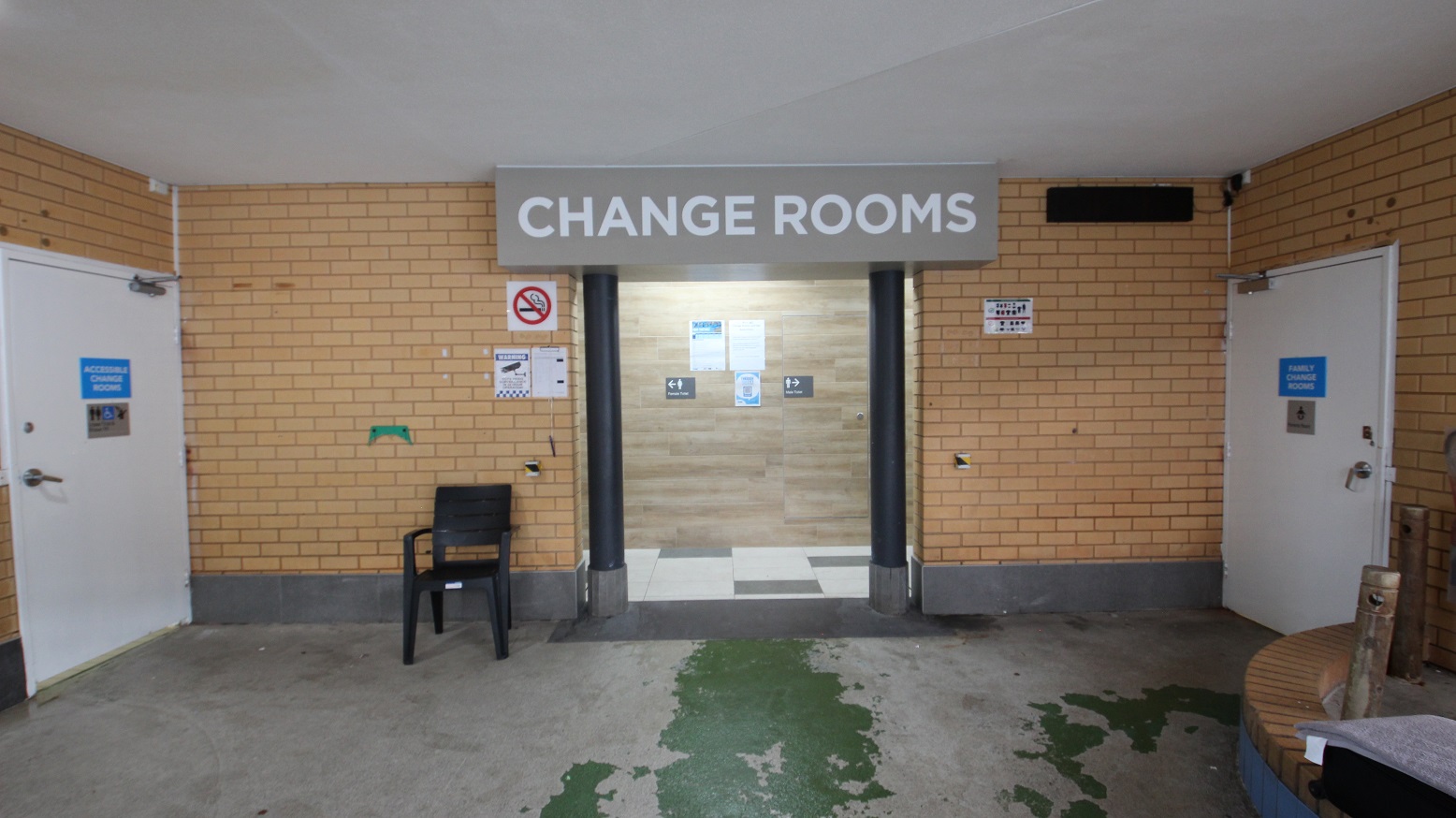 Change rooms at Whitlam Leisure Centre can be distinguished by the large brown block with black posts at the entrance. The ceiling is white, while the walls are made of bricks. Two white accessible change rooms opposite each other can be utilized by persons with mobility disabilities. A smoking sign and a CCTV sign are placed on the brick wall