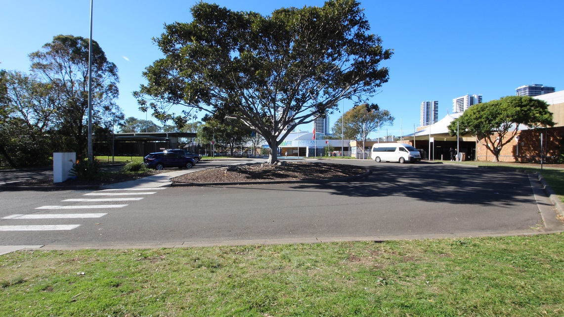 A white HI-ACE van is parked in one of the accessible car parks at Whitlam Leisure Centre, as marked by the accessible parking sign painted in blue and white. On its left side is an accessible parking sign only for persons with a mobility disability and an arrow pointing in the direction of the van. Two yellow bollards are erected in the space connected to the pathway leading to a pedestrian lane.