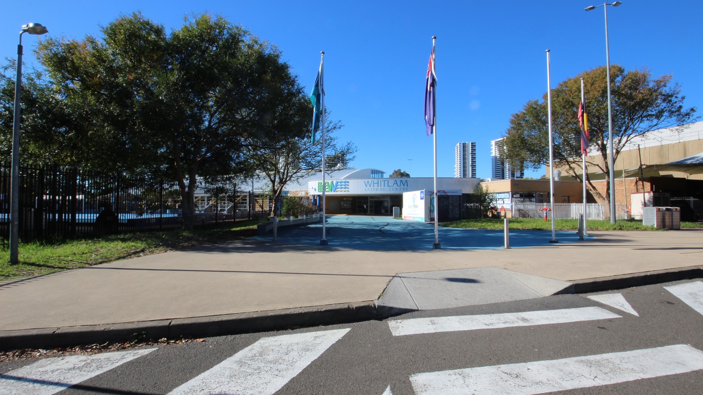 A zebra crossing leading to the smooth, concrete, blue pathway to the entrance of Whitlam Leisure Centre. At the boundary of the grey pathway are flag poles, flags, and grey bollards. There are also lamp posts assigned to opposite sides of this wide pathway. A large tree inside the black fence and pool provides great shade to the pathway.