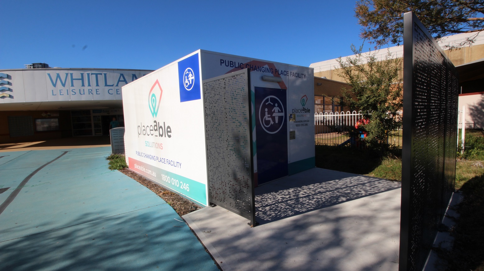 A closer look at the public changing place facility by Placeable Solutions. The right side of the black gate is opened and a square, dirty white, concrete pathway is placed in front of the facility. The facility has a dark blue door with accessible icons.