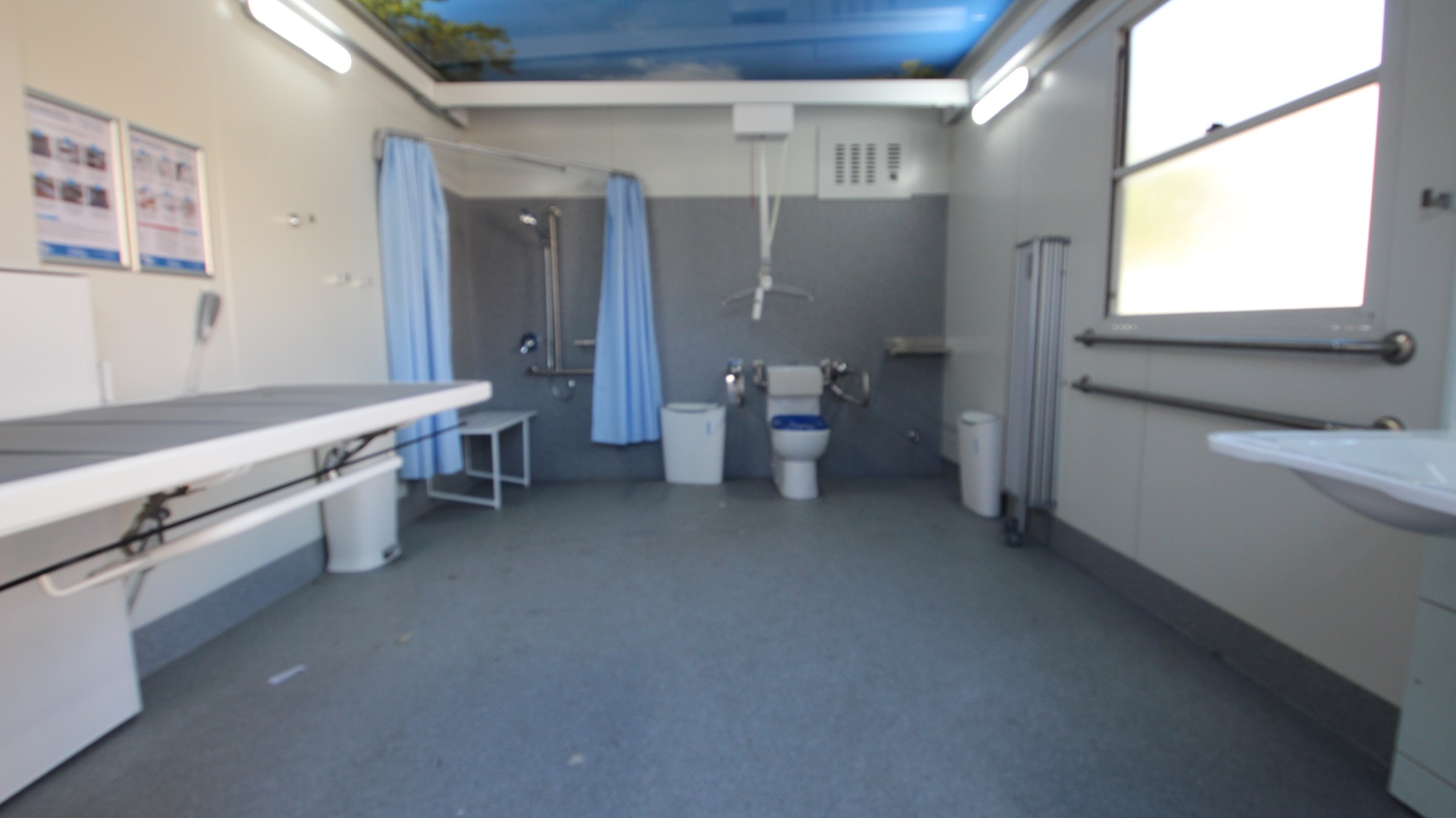 Inside the accessible bathroom at Whitlam Leisure Centre is a hoist, a dark blue cushioned adult change table, a toilet, and a shower with a sky blue privacy curtain. Beside the toilet bowl is a white rubbish bin. The back wall is painted grey, while the left and right are smooth white walls. Near the entrance, to the left, is a wash basin.