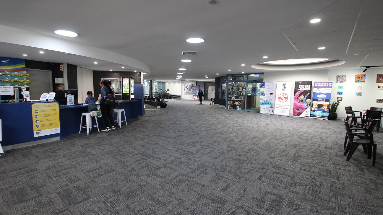 Next to the curvy reception tables is a thigh-light counter table with tall white bar stools. Heading towards the right from the reception desk are the Health Club and Swell Café. Different wall paintings and posters displayed near the wall can be seen opposite the reception area as well as black chairs and tables.