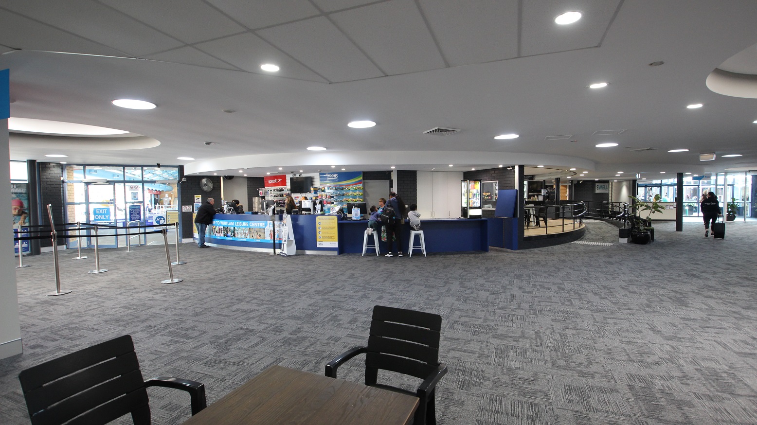 View when standing in the seating area containing black chairs and tables opposite of the reception desk. This image includes the peripheral part of the Swell Café with a low ramp-style entry point. The ramp has black metal handrails, and against it is a medium-sized potted plant and black chairs.
